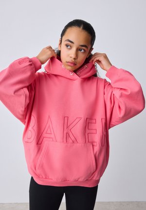 Jeune personne ajustant la capuche d’un sweat à capuche oversize rose vif avec un grand texte « BAKER », debout devant un fond uni clair.
