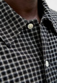 Primer plano de un cuello de camisa a cuadros en blanco y negro con dos botones blancos abrochados en el cuello y la parte superior del pecho de una persona.