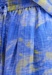 Tkanina plave i žute boje s uzorkom tie-dye i naboranom teksturom; ima elastičnu traku u struku i nabrani dizajn. Lagani i lepršavi materijal.