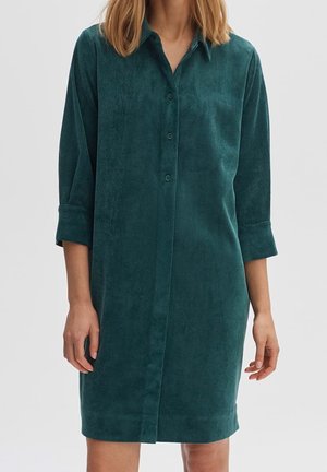 Femme portant une robe chemise boutonnée verte foncée à manches longues avec col, debout devant un fond clair uni.