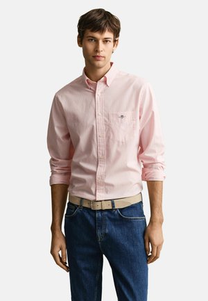 Mann trägt ein hellrosa Hemd mit hochgekrempelten Ärmeln, einen beigen Gürtel und blaue Jeans vor einem einfachen Hintergrund.