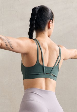 Grüner Sport-BH mit mehreren dünnen Trägern, aus einem glatten Stoff gefertigt. Mit ausgeschnittenen Details und einem kleinen Nike-Logo. Kombiniert mit helllila Leggings.
