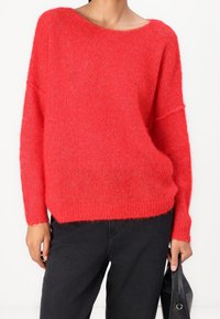 Pull rouge en tricot avec une coupe ample, un col large et des manches longues, doté d'une texture douce et d'un ourlet côtelé, porté avec un pantalon noir.