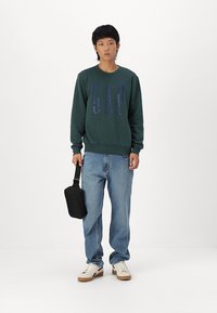 Hanorac de culoare verde închis cu logo-ul "GAP" în albastru mai închis, asortat cu blugi din denim deschis și adidași albi. Poartă o geantă neagră pe umăr.