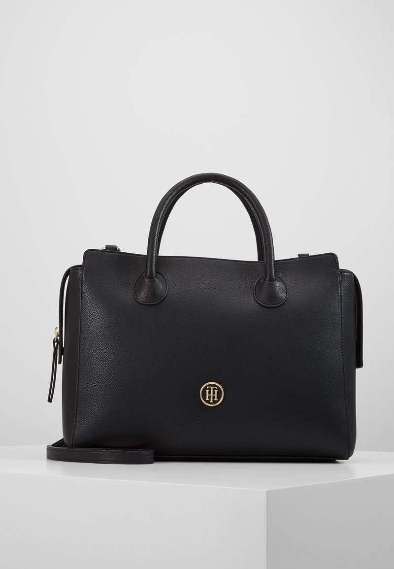 Sac à main en cuir noir avec double anses, une bandoulière amovible, et un petit logo rond en or sur le devant.