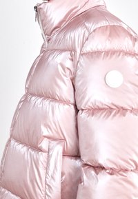 Lyserød dunjakke, skinnende materiale, quiltet design, høj krave, sidelomme, cirkulært hvidt mærke på ærmet.