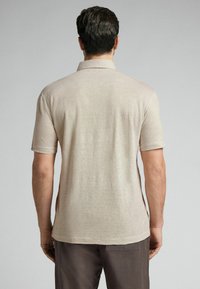 Beige polo tričko s krátkým rukávem a klasickým límečkem, měkkou texturou a bočními rozparky. Pohled zezadu, s lehkým materiálem splývajícím přes ramena.