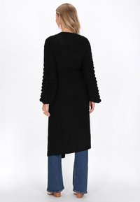 Dlouhý černý pletený cardigan s texturovanými rukávy, stahovaným pasem, otevřeným předním dílem a střední délkou, sladěný s širokými modrými džíny.