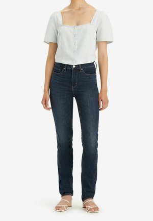 Femme portant un chemisier blanc à manches courtes et à col carré, un jean slim bleu foncé et des sandales blanches à talons, debout devant un fond blanc.