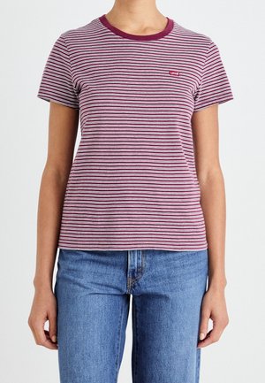 Person iført en kortærmet vinrød og hvid stribet t-shirt med et lille Levi's-logo, kombineret med blå jeans mod en ensfarvet baggrund.