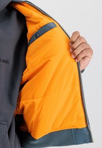 Bunda na zips s šedým vonkajším materiálom a jasne oranžovou podšívkou. Obsahuje kontrastný námornícky akcent na rukáve. Štruktúrovaná tkanina.