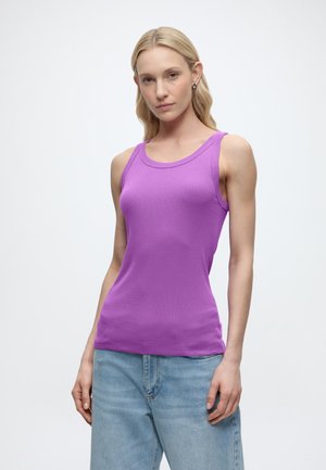 Γυναίκα με μακριά ξανθά μαλλιά, που φοράει μωβ αμάνικο τοπ και ανοιχτόχρωμο τζιν, στέκεται μπροστά σε λευκό φόντο.