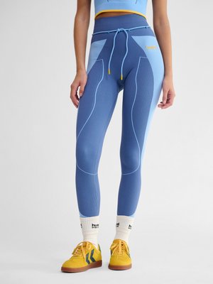 Frau trägt blaue nahtlose Leggings mit Kordelzug, weiße Sportsocken und gelbe Sneakers, steht vor einem einfarbigen Hintergrund.