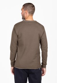 Camicia a maniche lunghe in cotone verde oliva, con scollatura rotonda e orli rifiniti, indossata con pantaloni blu navy. Il tessuto appare morbido e liscio.