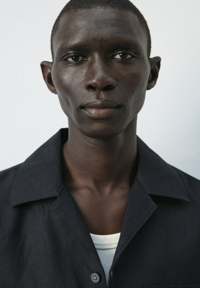 Gros plan sur un jeune homme noir aux cheveux courts portant une chemise à col foncée sur un haut blanc, regardant droit devant avec une expression neutre.