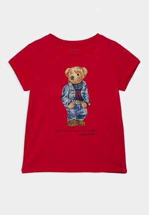 Czerwona koszulka z krótkim rękawem z grafiką misia w ubraniu: kurtka jeansowa, jeansy i buty, stojącego z rękami w kieszeniach.