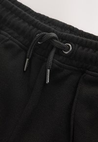 Gros plan sur un pantalon en tissu noir texturé avec un cordon noué passé dans des œillets métalliques à la ceinture.