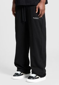 Black sweatpants with a relaxed fit, featuring side pockets and a logo in white. Paired with black and white sneakers.