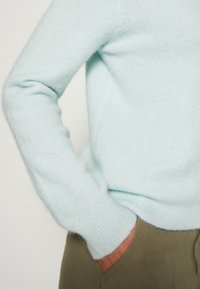 Suéter de color azul claro, suave, con cuello redondo y mangas largas, que presenta puños acanalados. La mano está metida en un bolsillo de unos pantalones verde oliva.