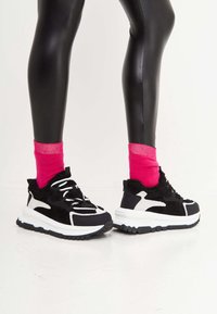 Jambes en leggings noirs brillants portant des baskets épaisses noires et blanches avec des chaussettes rose vif sur un sol et un fond blancs.