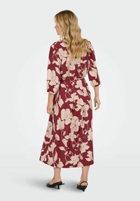 Lång vinröd klänning med krämfärgad blommönster, trekvartslånga ärmar, bälte i midjan och en flödande siluett. Buren med svarta slingback-skor.