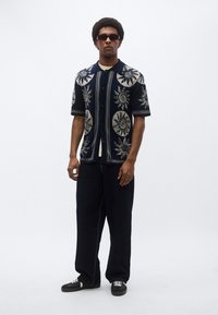 Μπλε ναυτικό πουκάμισο με κοντά μανίκια και σχέδια ήλιου, κουμπιά και γιακά, συνδυασμένο με σκούρο τζιν παντελόνι και μαύρα αθλητικά παπούτσια. Αξεσουάρ περιλαμβάνουν γυαλιά ηλίου.