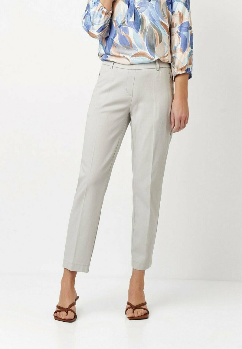 Helle graue Hosen mit schmal zulaufenden Beinen, flacher Vorderseite und Gürtelschlaufen, kombiniert mit einer bunten, blumengemusterten Bluse und braunen, geschnürten Sandalen.