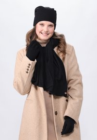 Berretto di maglia nero, sciarpa nera abbinata e guanti indossati con un cappotto beige. La sciarpa ha una superficie liscia e un accento di marca.