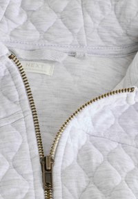 Veste matelassée blanche à zip, avec une fermeture éclair dorée et une étiquette en tissu, mettant en avant un motif texturé dans une palette de couleurs claires.