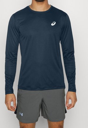 Mann trägt ein marineblaues langärmeliges Sportshirt und graue Shorts, steht mit entspannt herabhängenden Armen an den Seiten vor einem einfarbigen Hintergrund.