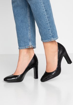 Femme portant un jean bleu et des chaussures à talons hauts noires texturées, debout sur un sol gris clair devant un fond uni.