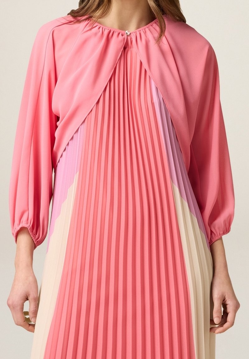 Femme portant une robe plissée rose avec de longues manches bouffantes et un décolleté en trou de serrure fermé par un petit bouton.