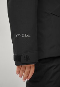 De waterdichte zwarte jas heeft manchetkenmerken met het witte geborduurde "67°N áhkka" logo en een gestructureerde zwarte klittenbandsluiting met geometrisch patroon detail.