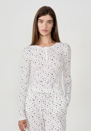 Woman with long brown hair wearing white ribbed pajama set with pink and black heart pattern, standing against plain light background.