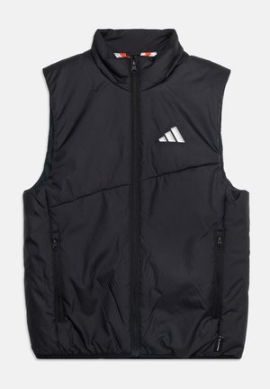 Barn iført en sort dunvest, sort langærmet skjorte, grønne patchwork-bukser og sorte sneakers, der holder en hvid vandflaske.