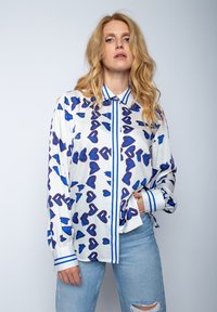 Biała koszula z długim rękawem w niebieski wzór serc, z kołnierzykiem na guziki, pionowymi niebieskimi paskami na froncie i mankietami z podwiniętymi rękawami.