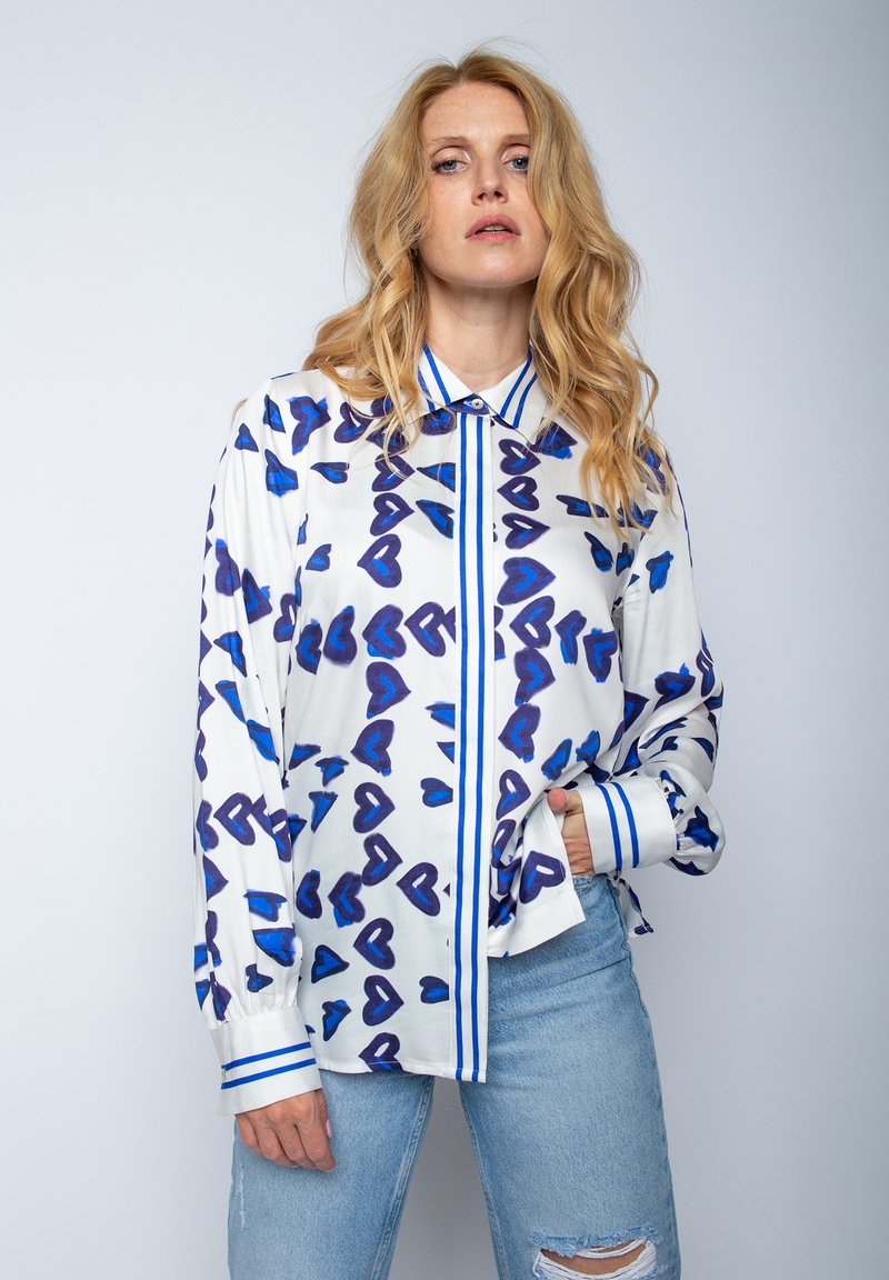 Biała koszula z długim rękawem w niebieski wzór serc, z kołnierzykiem na guziki, pionowymi niebieskimi paskami na froncie i mankietami z podwiniętymi rękawami.