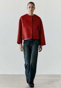 Giacca corta in lana rossa con scollo rotondo e bottoni ton sur ton, abbinata a jeans denim scuri. Tessuto liscio, vestibilità rilassata, su sfondo neutro.