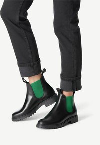 Bottes basses en cuir noir avec une semelle texturée, dotées de panneaux latéraux élastiques verts et de languettes de tirage, portées avec un pantalon en denim noir retroussé.