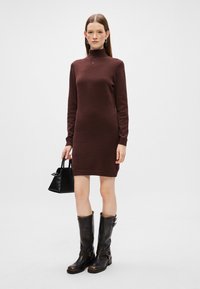 Vestido marrón de punto de manga larga con cuello alto, combinado con un bolso negro y botas altas de cuero marrón. Diseño sencillo, sin patrones.