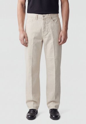 Uomo che indossa pantaloni beige a gamba dritta, cintura nera, scarpe nere e una camicia nera, in piedi davanti a uno sfondo chiaro e uniforme.