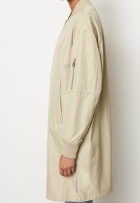 Beige bomberjack met ribbelmanchetten, zijzakken en een ritszak op de mouw, gemaakt van een gladde, lichte stof.