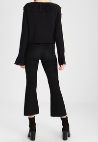 Blouse noire courte avec col à volants, associée à un jean noir évasé. Tissu texturé sur le pantalon ; bottines à la cheville complètent le look.