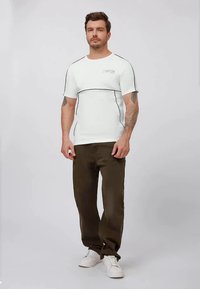 Man staan in een wit T-shirt met zwarte accenten, bruine broeken, witte sneakers en zichtbare arm tattoos voor een effen achtergrond.