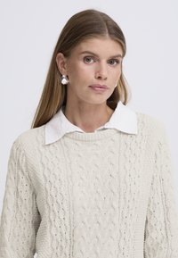 Maglione crema lavorato a maglia con motivo a trecce, abbinato a una camicia bianca con colletto. Presenta orecchini a goccia d'argento, texture liscia e vestibilità comoda.