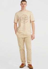 Jonge man die met handen in zijn zakken staat, gekleed in een beige graphic T-shirt, beige broek en bruine sneakers, tegen een witte achtergrond.