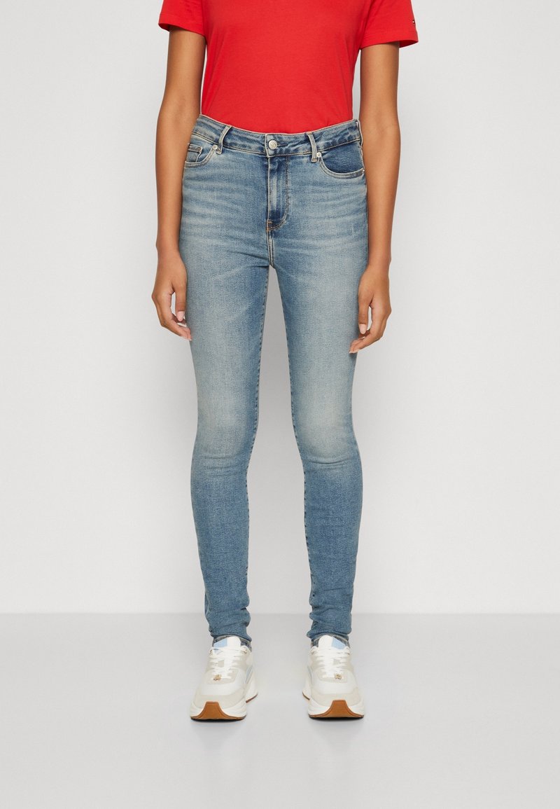 Jeans ajustados de tiro alto en azul claro. Presentan bolsillos delanteros, un ligero desvanecimiento y costuras en contraste. Combinados con una camiseta roja y zapatillas blancas.