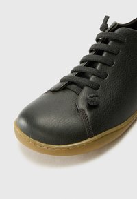 Scarpa in pelle nera con punta arrotondata, tomaia texture, lacci grigi e suola in gomma marrone chiaro. Presenta un accento a forma di anello vicino alla linguetta.