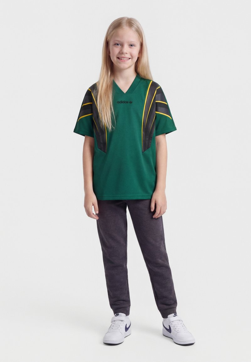 Enfant souriant aux longs cheveux blonds, portant un maillot de sport vert Adidas, un pantalon de jogging foncé et des baskets blanches Nike, debout sur un fond blanc.