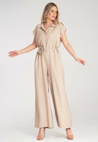 Beżowy kombinezon z krótkimi rękawami, zapinany na guziki z materiałowym paskiem z detalem w postaci kwiatu. Szerokie nogawki, gładka faktura i lekki materiał.